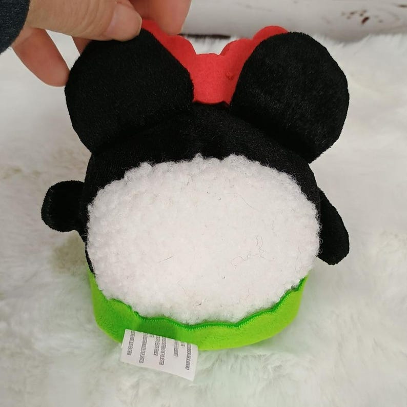 Pu&ograve; includere: Un peluche a forma di testa di personaggio, con orecchie nere, un fiocco rosso e un corpo bianco e soffice. La base &egrave; verde. Il giocattolo &egrave; tenuto in mano.