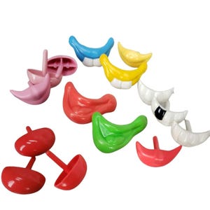 Può includere: Una collezione di bocche e labbra giocattolo in plastica colorata in varie tonalità di rosso, rosa, verde, giallo, blu e bianco. Alcuni pezzi hanno steli, altri denti. I giocattoli sono sparsi su uno sfondo bianco.