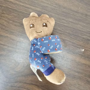 May include: A plush toy shaped like a tree character with a light brown head and body. The toy is wearing blue fabric clothing featuring a repeating pattern of small white trees and red mushrooms, with a dark blue band at the bottom of the leg.