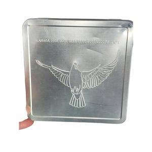 Puede incluir: Una lata de metal plateada con un diseño de paloma y el texto "CANADA 1998 1999 1999 1999 2000 2000 2000 2000 CANADA".