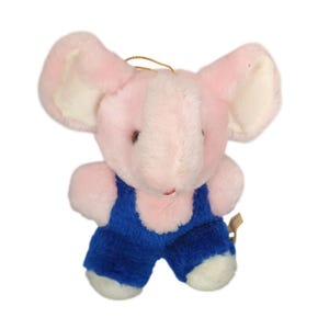 Könnte beinhalten: Ein kleines, rosa Plüschelefanten-Spielzeug mit großen Ohren und einer blauen Jeans-Latzhose. Der Elefant hat weiße Akzente an Ohren und Füßen. Zum Aufhängen ist eine goldene Schnur befestigt. Das Spielzeug ist für Kinder konzipiert.