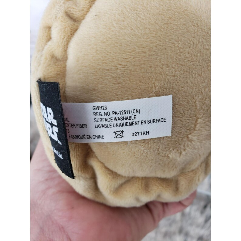 Op de afbeelding: Een bruin pluche speelgoed met een label met de tekst "STAR WARS" en "GWH23 REG. NO. PA-12511 (CN) SURFACE WASHABLE LAVABLE UNIQUEMENT EN SURFACE FABRIQU&Eacute; EN CHINE". Het speelgoed is gemaakt van polyestervezel en is oppervlakte wasbaar.