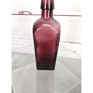 May include: A vintage, deep purple glass bottle with a rectangular base and a long neck. The bottle has a rounded shoulder and a label design. The text "VASE-LINE" and "SINCE 1882" are embossed on the bottle.