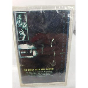 May include: A vintage cassette tape of the album "Snap!" in a clear plastic case. The album cover features the band's logo and the text "The Debut With Real Power!" in white. The background is dark with a blue and white design.