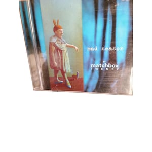 May include: CD cover art for the album "Mad Season" by Matchbox Twenty. The cover features a cartoon-like illustration of a person wearing a pink bunny costume and a blue dress, standing in front of a blue and white background.