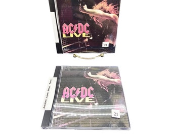 Puede incluir: Una caja de CD negra con una portada de álbum AC/DC Live rosa y blanca. La portada del álbum presenta una foto de una persona tocando la guitarra. El texto "AC/DC Live" está impreso en rosa y blanco en la portada. El número "21" está impreso en blanco en la esquina inferior derecha de la portada.