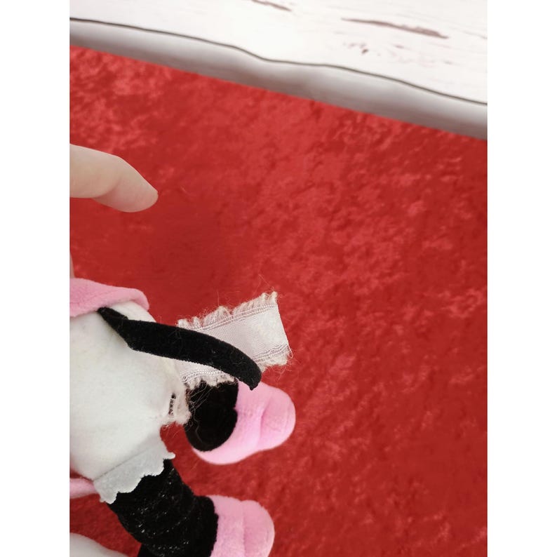 May include: A soft toy cow featuring a pink hat, a black and white body, and pink feet. The toy is positioned against a red backdrop. The cow's black and white body is visible, along with its pink feet and hat.