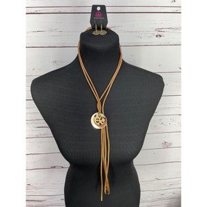 May include: A brown leather cord necklace with a gold pendant featuring a leopard print design. The necklace is displayed on a black mannequin. Matching earrings are included.