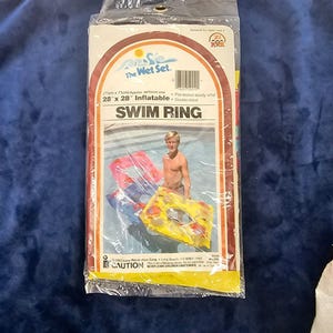 Può includere: Una confezione in plastica trasparente contenente un salvagente gonfiabile da 71 cm per 71 cm. La confezione riporta la scritta The Wet Set, SWIM RING e l'illustrazione di una persona in piscina con un salvagente giallo e rosso a motivi.