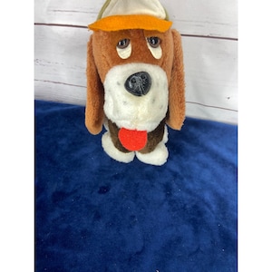 Könnte beinhalten: Ein Vintage-Plüschhundespielzeug mit braunen, weißen und orangefarbenen Akzenten. Der Hund hat Schlappohren, eine schwarze Nase und eine rote Zunge. Er trägt einen Hut mit grünem und orangefarbenem Rand.