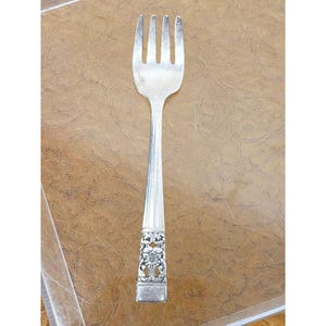 May include: A silver-toned metal fork featuring four tines and a smooth, tapered handle. The lower portion of the handle is decorated with an intricate, raised floral relief pattern, while the upper section remains plain and polished.