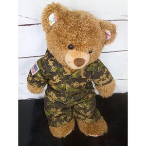 May include: A brown plush teddy bear wearing a military-style uniform with a green and brown digital camouflage pattern. The uniform features a small American flag patch on the right sleeve and the bear has colorful patterned fabric inside its ears.