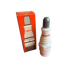 Può includere: Una bottiglia bianca a forma di candela Champion con tappo grigio e la scritta "Champion" in rosso. La bottiglia è accanto a una scatola arancione con una grafica di candela e la scritta "Champion".