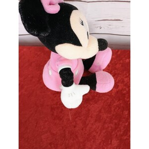 May include: A plush Minnie Mouse doll, with black fur, a pink bow, and a pink dress with white polka dots. The doll has white gloves and pink shoes. The doll is sitting on a red surface.