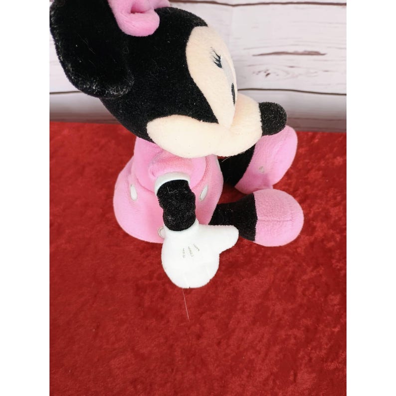 May include: A plush Minnie Mouse doll, with black fur, a pink bow, and a pink dress with white polka dots. The doll has white gloves and pink shoes. The doll is sitting on a red surface.