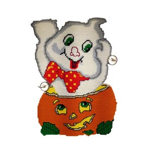Puede incluir: Decoración de Halloween hecha a mano con un fantasma sonriente que emerge de una calabaza naranja. El fantasma es blanco con una pajarita roja y ojos verdes. La calabaza tiene una cara tallada y detalles verdes.