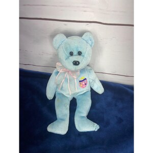 Peut inclure: Un ours en peluche bleu clair Beanie Baby avec un ruban rose autour du cou et un patch d'œuf de Pâques sur la poitrine. L'ours est debout sur une surface bleue.