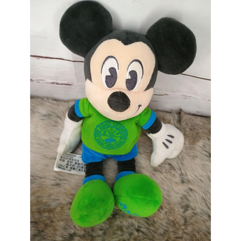 May include: A plush Mickey Mouse toy, featuring a black head and ears, a tan face, and white gloves. Mickey wears a green shirt with a blue emblem, blue shorts, and green shoes. The toy is on a light-colored surface.