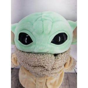 Op de afbeelding: Een pluche speelgoed van een personage met grote zwarte ogen, grote oren en een lichtgroene teint. Het personage draagt een bruine jas met een fuzzy kraag. Het speelgoed is een populair personage uit een sciencefictionreeks.
