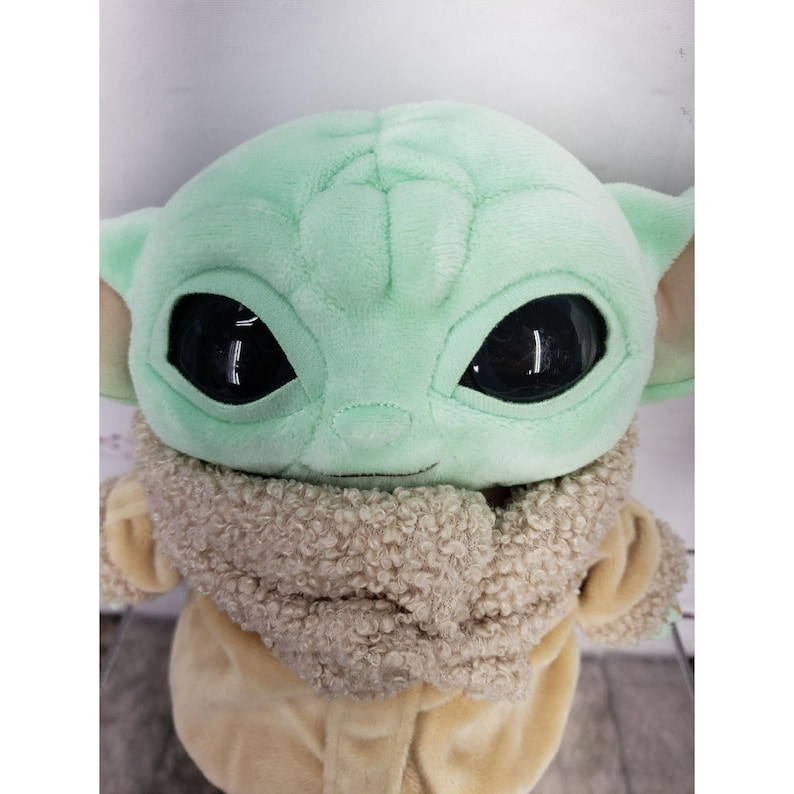 Op de afbeelding: Een pluche speelgoed van een personage met grote zwarte ogen, grote oren en een lichtgroene teint. Het personage draagt een bruine jas met een fuzzy kraag. Het speelgoed is een populair personage uit een sciencefictionreeks.