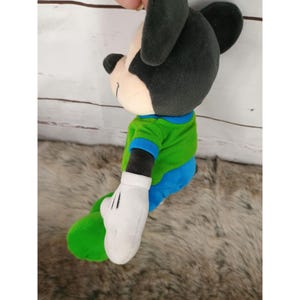 May include: A plush Mickey Mouse toy in a seated position. The toy has a black head and ears, a cream-colored face, and a green shirt with blue trim. The shorts are blue, and the hands are white with black lines. The toy is on a light brown surface.