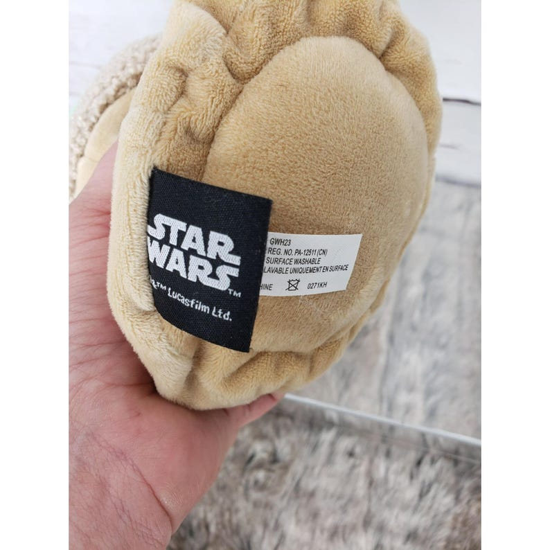 Op de afbeelding: Close-up van de voet van een pluche speelgoed, bruin van kleur met een getextureerd oppervlak. Een zwart label toont het "STAR WARS" logo en tekst die aangeeft dat het oppervlak wasbaar is. Het label bevat ook het handelsmerk Lucasfilm Ltd.