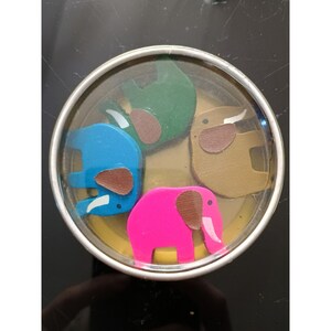 May include: A clear, round container holds four colorful wooden elephant figurines. The elephants are blue, green, brown, and pink, each with a brown ear and white tusk. The container has a silver rim.