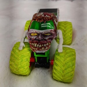 May include: A toy monster truck with a zombie-themed design. The truck features a green chassis, large lime green tires, and a detailed zombie head with exposed teeth and blood-red accents. The truck has a black top.