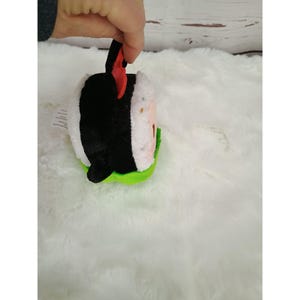 Pu&ograve; includere: Un giocattolo di peluche a forma di rotolo di sushi con alghe nere, riso bianco e una guarnizione rossa. Il giocattolo ha una base verde ed &egrave; tenuto in mano. Il giocattolo &egrave; su una superficie bianca e soffice.