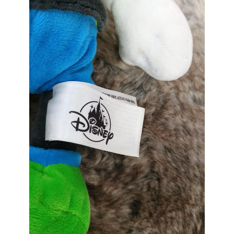 May include: A plush toy with blue, green, and white sections. A white tag with the Disney logo and text is attached to the toy. The background is a fuzzy, brown surface.