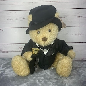 Peut inclure: Un ours en peluche portant un smoking noir et un chapeau haut de forme, tenant une trompette dorée. L'ours est assis sur une surface grise. La trompette a un ruban noir avec l'année "2000" imprimée dessus.