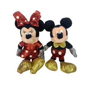 以下が含まれることがあります： 2体のディズニーキャラクターのぬいぐるみ、ミッキーとミニーマウスが並んで立っています。ミニーは白い水玉模様の赤いドレスとリボン、金色のグリッターシューズを履いています。ミッキーは赤い水玉模様のショーツ、金色の蝶ネクタイ、金色のグリッターシューズを履いています。