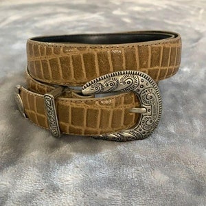 May include: A brown leather belt with a silver buckle featuring an intricate design. The belt has a crocodile-embossed pattern.
