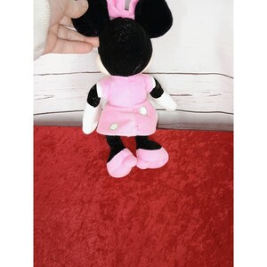 May include: A Minnie Mouse plush doll, with a black head and ears, a pink dress with white polka dots, and pink shoes. The doll has white gloves and is set against a red surface and a white wooden background.