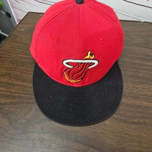 Op de afbeelding: Een rode honkbalpet met een zwarte klep en een geborduurd Miami Heat basketbal-logo op de voorzijde. Het logo toont een rood-gele vlammende basketbal met een witte omlijning. De pet heeft een zwarte knoop bovenop de bol van de pet.