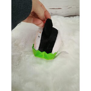 Pu&ograve; includere: Un onigiri (palla di riso) di peluche bianco, nero e rosa, con una base verde a forma di foglia. L'onigiri ha un involucro di alghe nere e un piccolo accento rosso. Il giocattolo &egrave; tenuto da una mano.