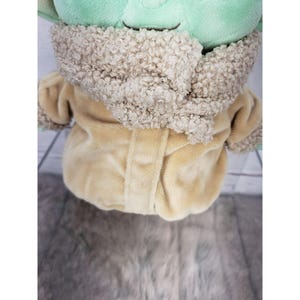 Op de afbeelding: Een pluche pop van een personage met grote oren en een groen gezicht, gekleed in een beige jas met een donzige, lichtbruine kraag. De pop zit op een lichtgrijze en witte houten ondergrond.