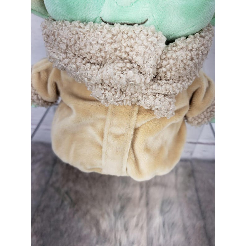 Op de afbeelding: Een pluche pop van een personage met grote oren en een groen gezicht, gekleed in een beige jas met een donzige, lichtbruine kraag. De pop zit op een lichtgrijze en witte houten ondergrond.
