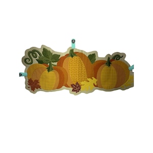Könnte beinhalten: Ein dekoratives Herbstbanner mit gestickten Kürbissen, Mais und Herbstblättern in Orange-, Gelb- und Brauntönen. Das Banner wird mit zwei hellblauen Clips an der Wand befestigt. Das Banner ist etwa 51 cm lang.
