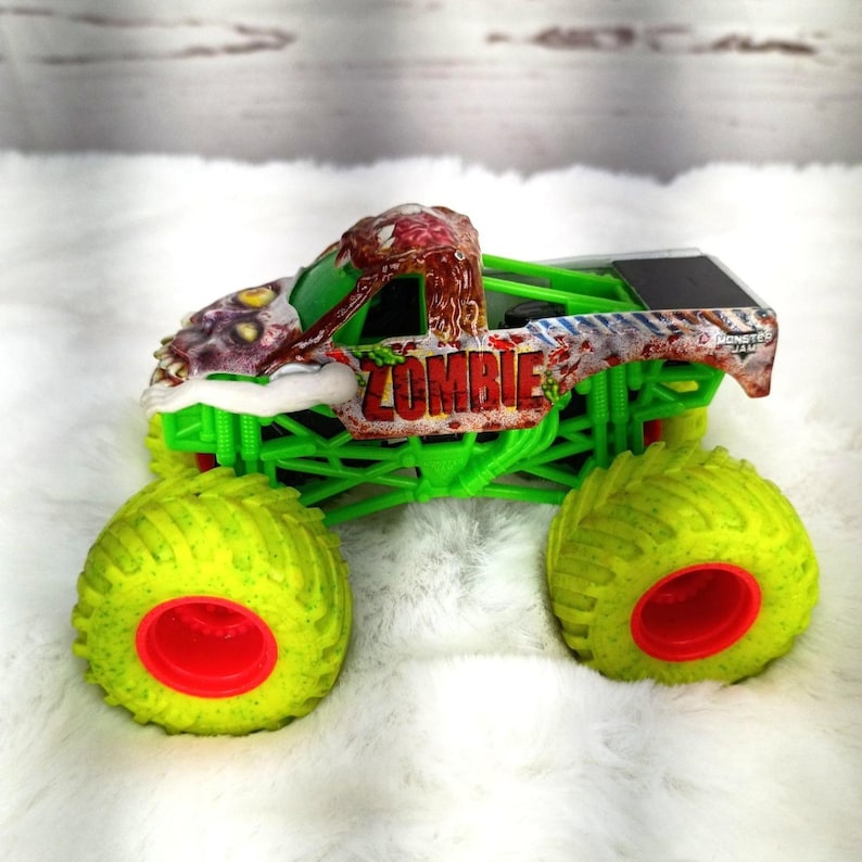 May include: A toy monster truck with a zombie theme. The truck features a green chassis, large yellow tires with red centers, and a cab with a zombie graphic and the word "ZOMBIE". The truck is on a white surface.