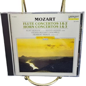 May include: A CD case with a yellow and orange sunset landscape on the front. The text on the front reads "Mozart Flute Concertos 1 & 2 Horn Concertos 1 & 3 Kurt Berger, Flute Bernd Heiser, Horn Vienna Mozart Ensemble Herbert Kraus, Conductor Digital Recording DDD Laserlight Digital 68 Min".