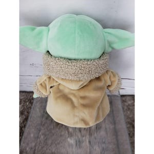 Op de afbeelding: Een pluche pop van een personage met grote oren en een lichtgroen hoofd. De pop draagt een bruine jas met een fuzzy kraag. De pop zit op een grijze ondergrond.