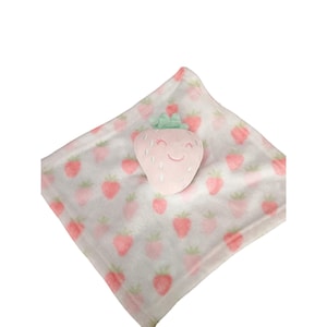May include: A baby blanket with a strawberry design. The blanket is white with pink strawberries and a pink strawberry plush toy with a smiling face. The strawberry plush has a green stem.