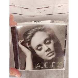 Puede incluir: Una caja de CD con una foto en blanco y negro de Adele en la parte delantera. El texto "ADELE 21" está impreso en blanco en la parte delantera de la caja.