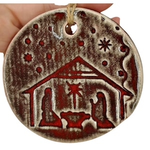 May include: A round, ceramic Christmas ornament with a nativity scene design. The ornament features a red and brown color scheme, with a depiction of the holy family, a star, and a stable. A jute string is attached for hanging.