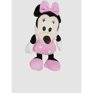 May include: A Minnie Mouse soft toy. The toy has a black head and ears, a white face, and a pink bow. She is wearing a pink dress with white polka dots, white gloves, and pink shoes. The doll is standing upright.