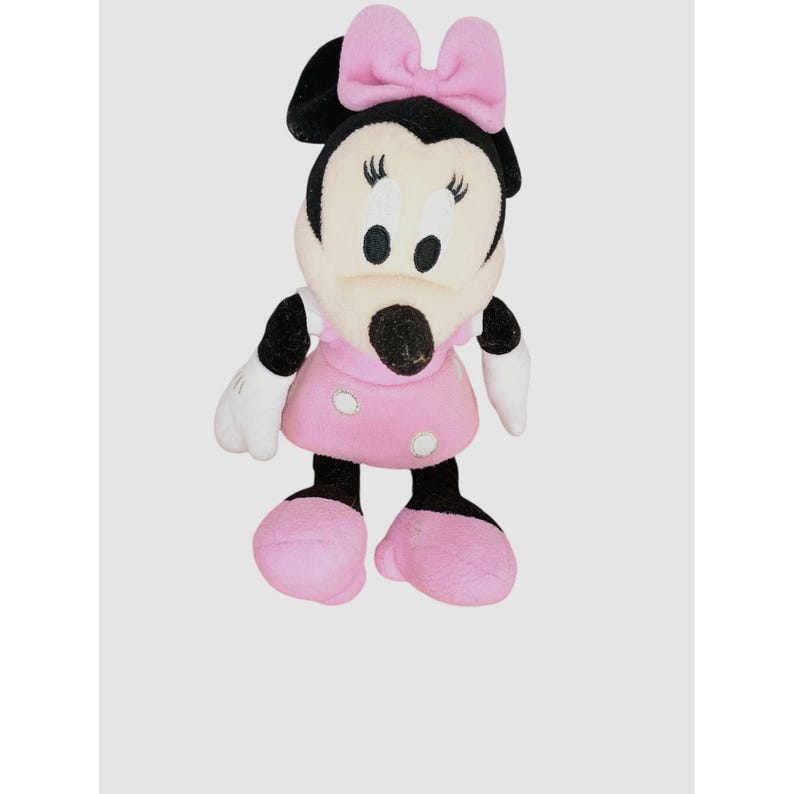 May include: A Minnie Mouse soft toy. The toy has a black head and ears, a white face, and a pink bow. She is wearing a pink dress with white polka dots, white gloves, and pink shoes. The doll is standing upright.
