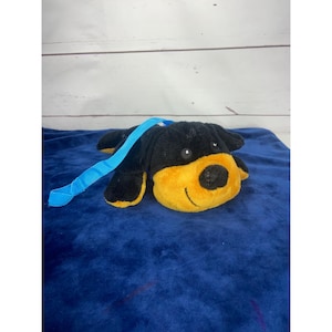 Op de afbeelding: Een zwarte en gele pluche hond met een blauwe band. De hond ligt met zijn hoofd naar de zijkant gedraaid.