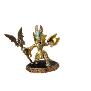 May include: A gold-coloured action figure of a character with an Egyptian pharaoh-inspired design. The figure is holding a staff with a wing-shaped top and has a teal-coloured necklace. It stands on a black base with a bronze-coloured border.