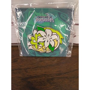 May include: A Creatures of Sonaria enamel pin featuring a stylized white flower with green leaves and yellow accents, attached to a branded backing card inside a clear plastic sleeve. The pin design includes a small green creature figure.